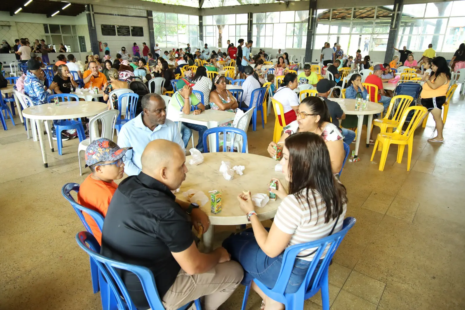 Foto de pessoas em um ambiente coberto, sentados em mesas, desfrutando de momentos de confraternização durante festa.