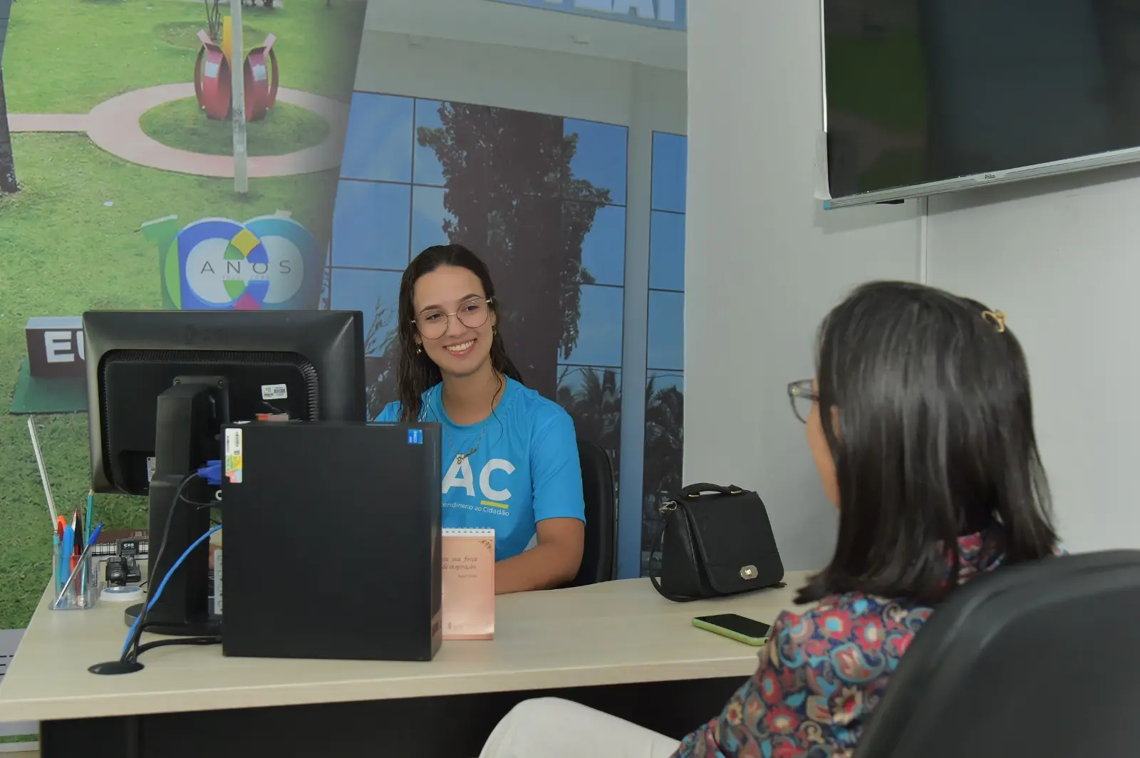 Foto de uma pessoa sentada a frente do computador atendendo outra a sua frente.