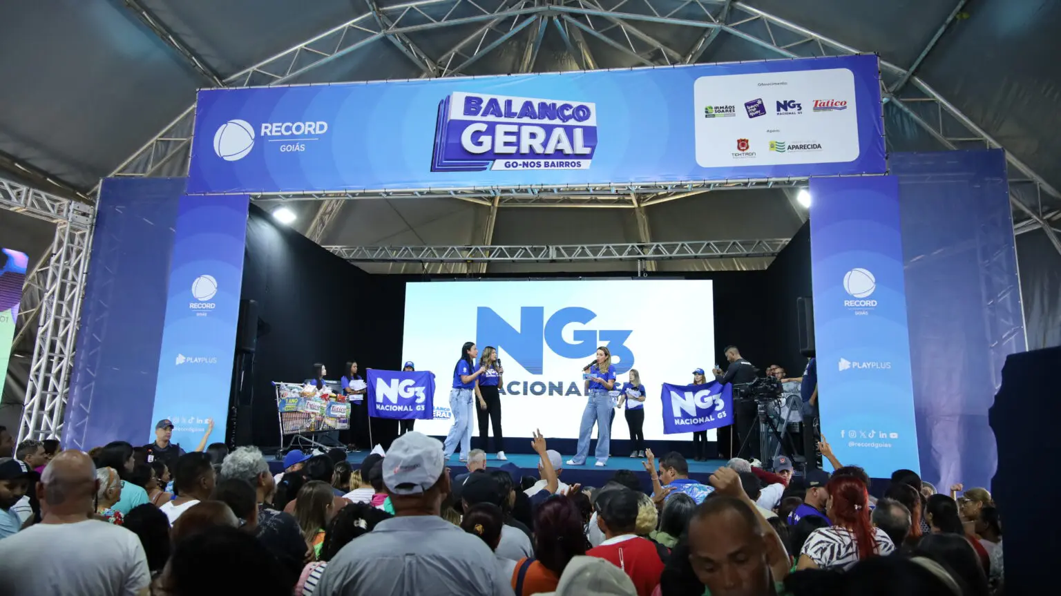 A foto mostra o palco principal de um evento do Balanço Geral nos Bairros, com apresentadores interagindo com o público. O espaço conta com grande estrutura e plateia.