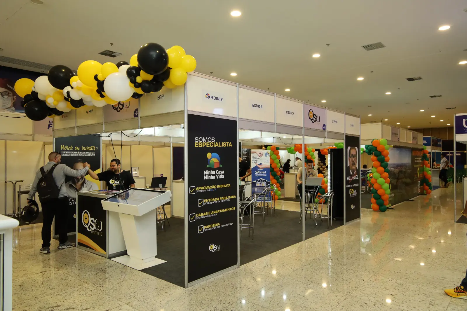 Foto do Feirão de Imóveis em Aparecida de Goiânia exibe estandes decorados e visitantes interagindo no evento.