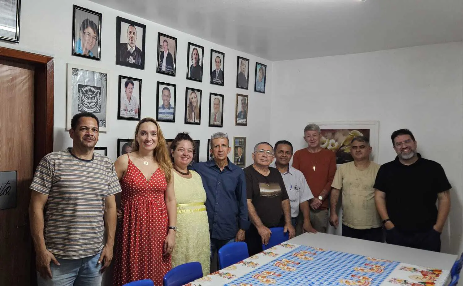 Membros de uma organização posam reunidos em um ambiente interno, com quadros de fotografias dispostos na parede ao fundo, destacando um momento de confraternização ou trabalho coletivo.