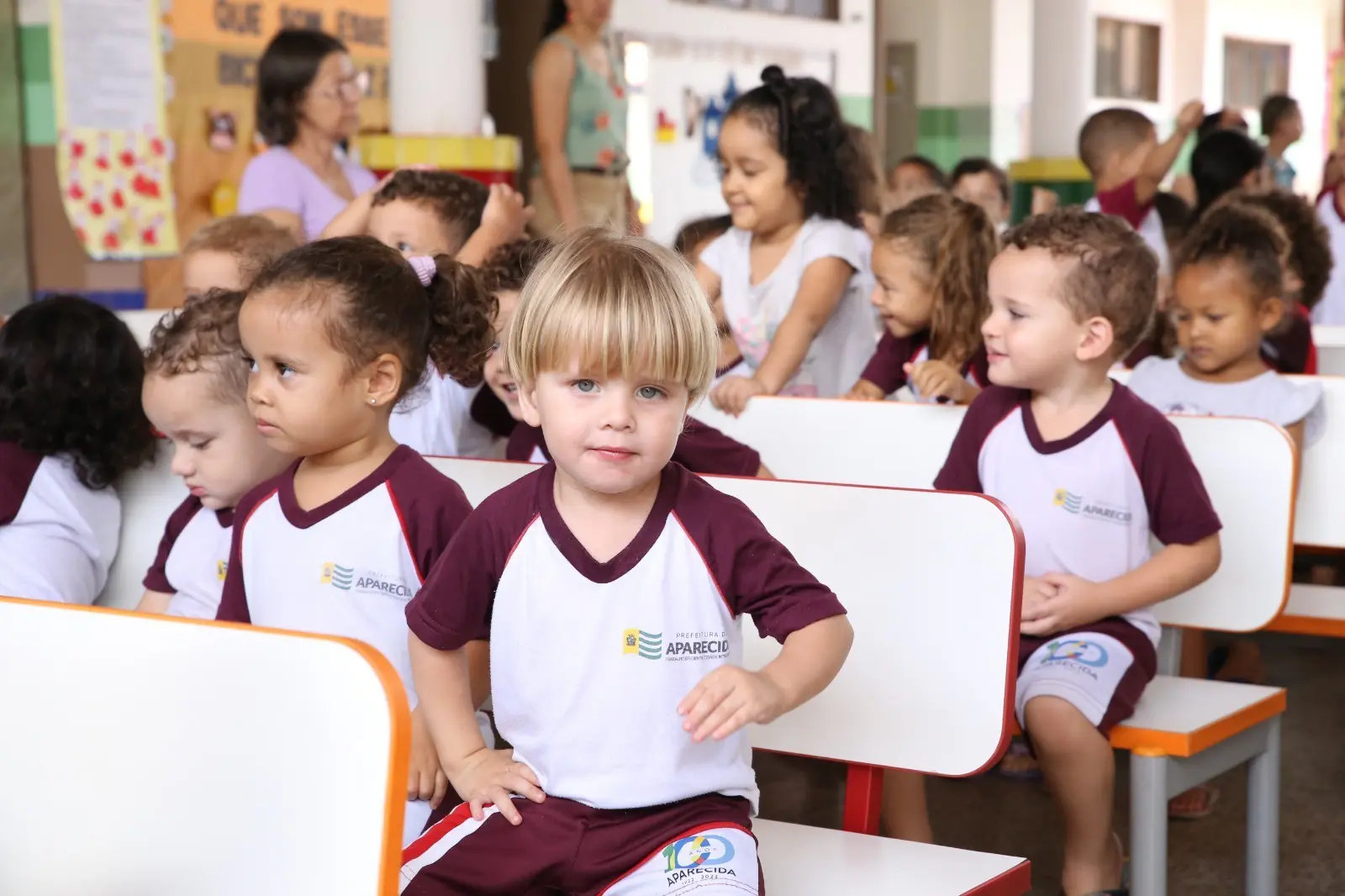 A foto mostra crianças uniformizadas, sentadas em cadeiras brancas com detalhes laranja, em um ambiente escolar do Cmei. Ao fundo, há murais decorativos, outras crianças e adultos interagindo.