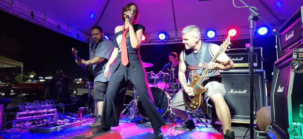 Foto de uma apresentação de uma banda de rock, tendo 3 pessoas portando seus instrumentos musicas em cima de um palco.