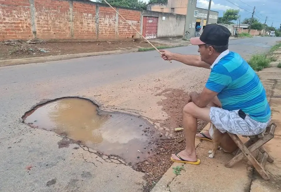 Na foto, um homem sentado em uma cadeira improvisada simula pesca com uma vara em um grande buraco cheio de água no asfalto de uma rua residencial.