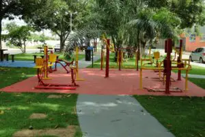 A foto mostra uma praça com equipamentos de academia ao ar livre em piso emborrachado, áreas gramadas, calçadas de concreto e bancos sombreados por árvores, destacando um ambiente urbano de lazer.