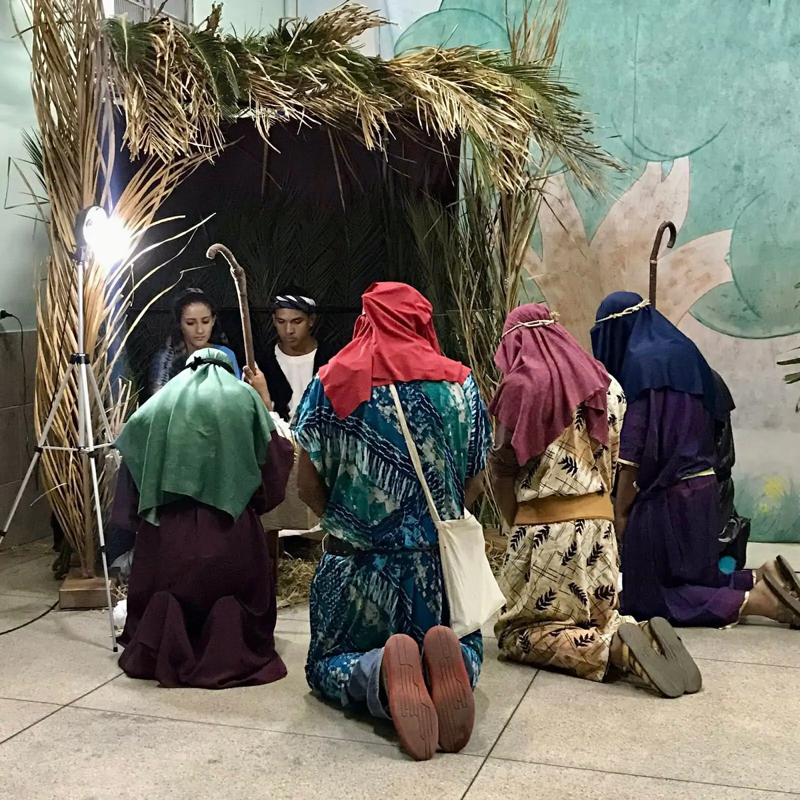 Foto de uma encenação do nascimento de Jesus, com Maria, José e pastores ajoelhados em um cenário decorado com palhas e folhas.