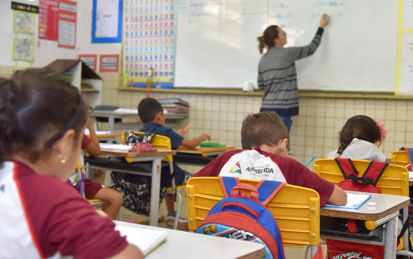 Crianças em sala de aula com uniformes da Prefeitura de Aparecida, realizando atividades enquanto a professora escreve no quadro branco.