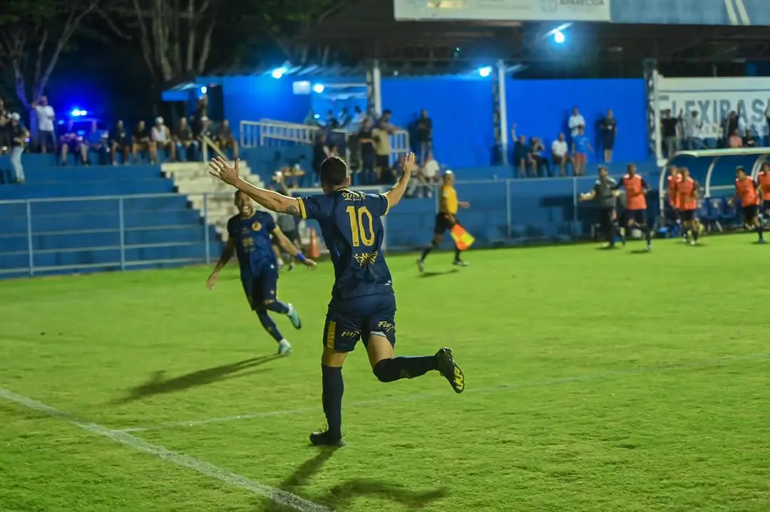 Jogador da Aparecidense celebra gol com braços abertos, correndo em direção à torcida, durante partida no Estádio Aníbal Toledo. Ao fundo, equipe e reservas comemoram.