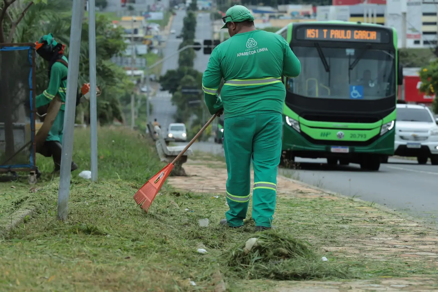 Trabalhadores realizam serviços de limpeza urbana, incluindo varrição e roçagem, em uma área pública movimentada com trânsito ao fundo.