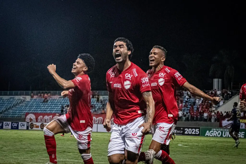 Jogadores comemoram intensamente em campo durante uma partida de futebol noturna, com uniformes vermelhos, sob luzes artificiais e diante de uma arquibancada parcialmente ocupada.