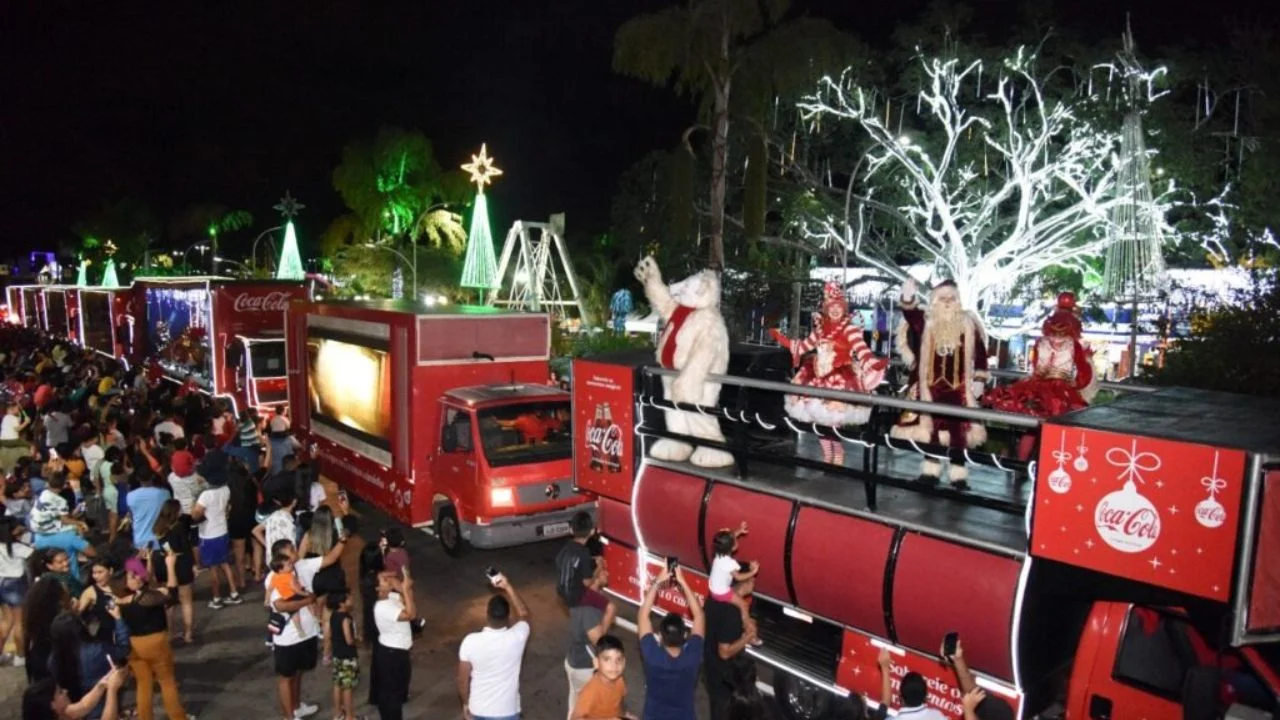 Foto de caminhões decorados com luzes natalinas e personagens temáticos encantam o público durante uma apresentação noturna, com destaque para a interação entre os espectadores e o Papai Noel.