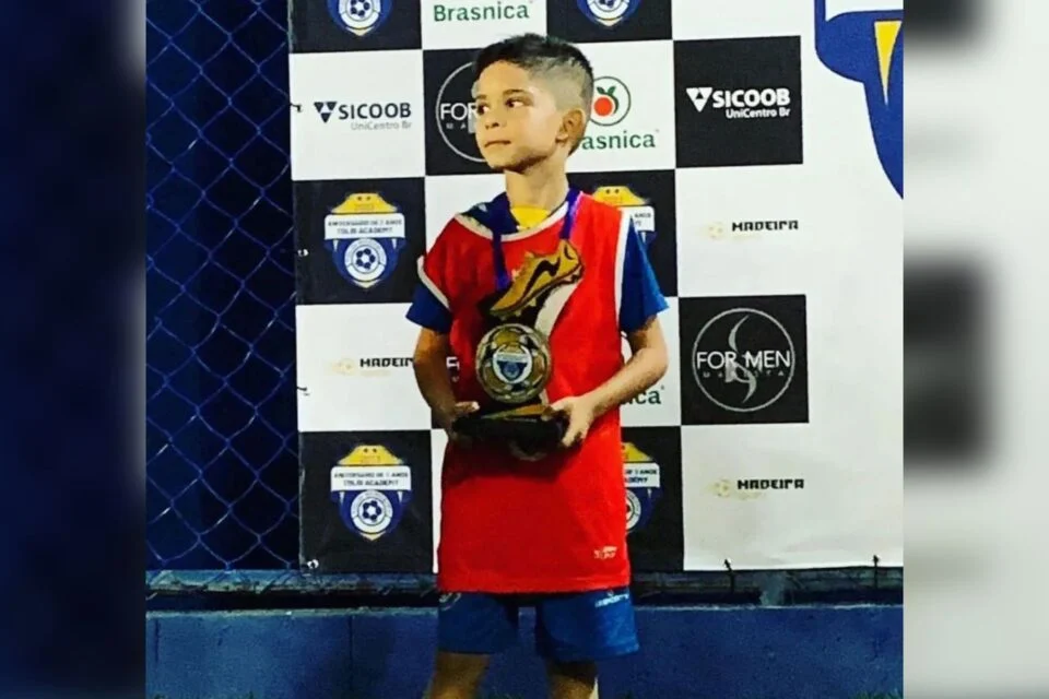 Foto de um garoto em uniforme de futsal segura um troféu com expressão confiante. Ele está em frente a um painel de patrocinadores, destacando sua conquista em uma competição.