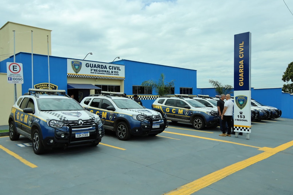 A imagem mostra a fachada da 3ª Base Regional da GCM, com viaturas estacionadas em frente, uma placa de identificação e duas pessoas conversando no local.