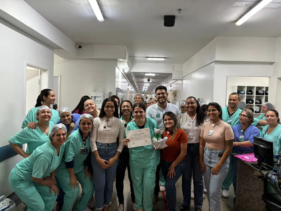 Profissionais de saúde posam juntos em um corredor de hospital, celebrando a entrega de um certificado de reconhecimento, em um momento de valorização e alegria coletiva.