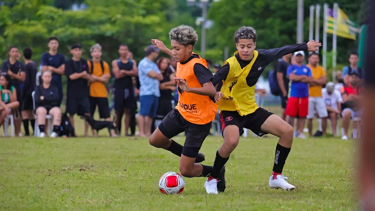 Dois jovens jogadores disputam intensamente a posse de bola em um gramado, sob o olhar atento de espectadores ao fundo.