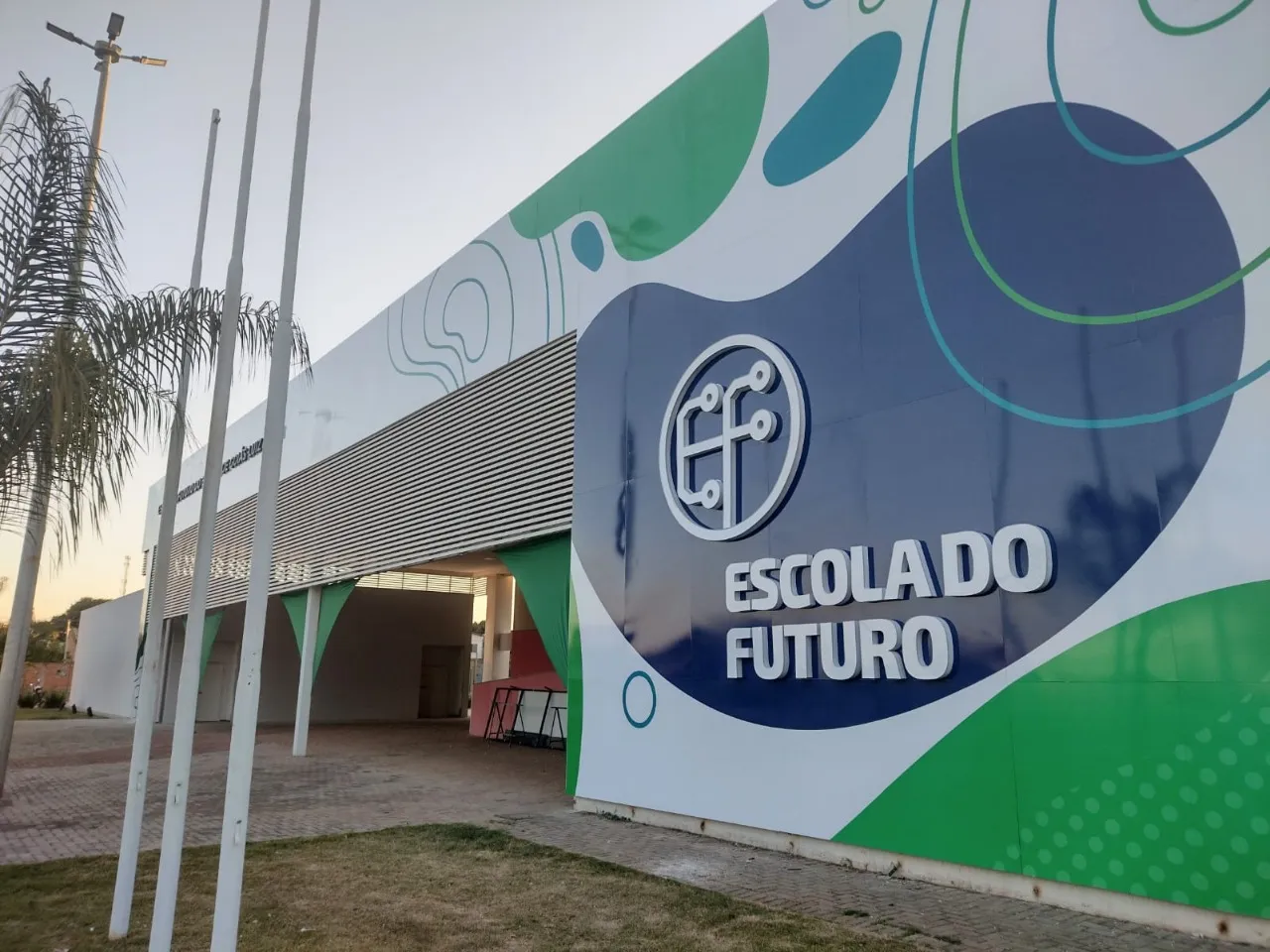 Fachada da unidade da Escola do Futuro, com design em verde e azul, destacando o logotipo da instituição e palmeiras ao lado, capturada ao entardecer.