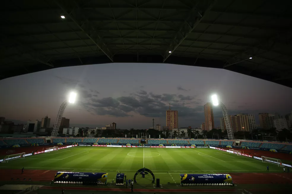 Estádio iluminado ao entardecer, com refletores acesos, arquibancadas vazias e uma paisagem urbana ao fundo, destacando prédios contra o céu em tons de azul e laranja.