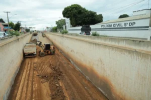 A foto registra obras de recuperação viária em um corredor de concreto, com máquinas removendo terra e resíduos, próximo à "Polícia Civil 5° DP"