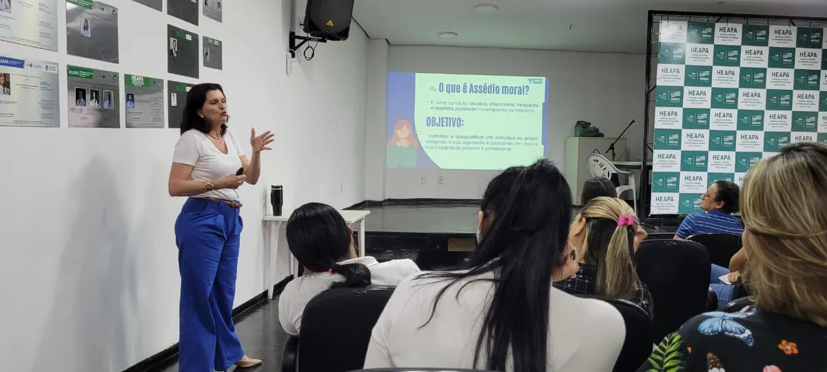 A imagem mostra uma palestra no Hospital Estadual de Aparecida de Goiânia (HEAPA) sobre assédio moral. A palestrante apresenta o tema em um telão, enquanto o público acompanha atentamente no auditório.